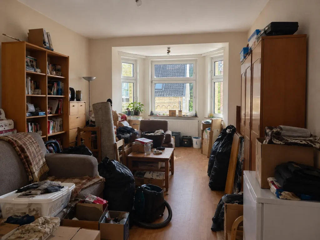 Vorher: ein überfülltes Wohnzimmer mit Erkerfenster, voller Möbel, Kartons, Müllsäcke und Wäschekörbe